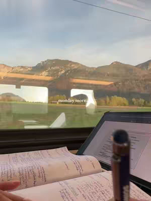 The image captures a study setup inside what appears to be a train or bus, with textbooks and a tablet open on a desk near a window. The window offers a view of mountains and a green landscape. Text overlayed says 'monday morning'.