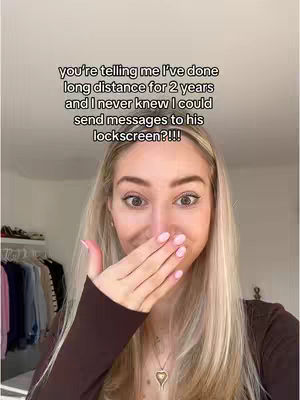 A selfie-style shot featuring a young woman with long blonde hair, covering her mouth with her hand. She's wearing a brown top and a necklace. The background has a soft blur, with hints of a clothing rack and a room.
