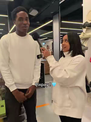 A man and a woman are featured facing forward and slightly toward each other in a retail environment. The man is on the left with dark hair wearing a Stone Island white sweatshirt and his hands are in front of him. The woman on the right is wearing white sweater and holding a microphone. The background suggests a store setting with shelves and overhead lighting. Below them, a text overlay reads 'Pixpay!' along with a blue heart.