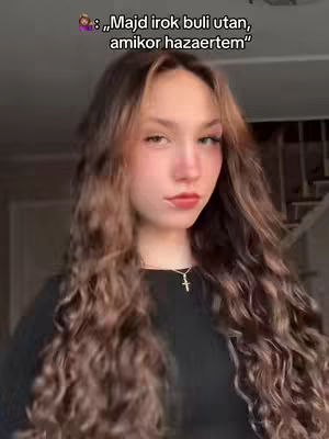 A young woman with long, wavy brown hair and red lipstick is centered in the frame. The background is softly blurred, likely a room with a staircase visible behind her. She has a necklace and a cross on it.