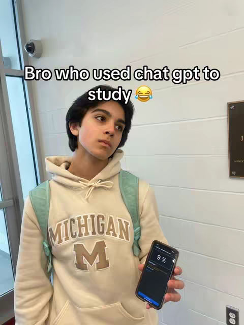A young man stands in a hallway, holding a phone. The background includes a white wall, a glass door, and a security camera. The man is wearing a cream-colored hoodie with a Michigan logo and a green backpack. The phone displays a 9% battery life notification. Text overlays the image.