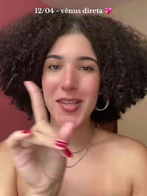 A woman with curly dark hair and fair skin is smiling and gesturing towards the viewer. She has a playful expression and is wearing earrings and a necklace. Text overlay is near the top left corner in white.