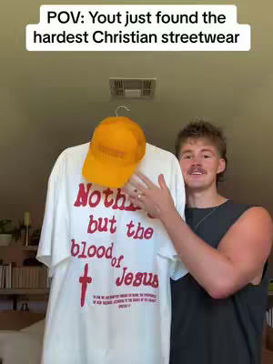 The image features a man holding up a white t-shirt and an orange cap. The t-shirt has red text that says "Nothing but the blood of Jesus" with a cross. The man is smiling, with a mustache, and dark hair. The wall in the background is beige.