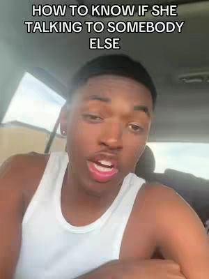 A young Black man is shown close up inside what appears to be a car. He is wearing a white tank top and speaking, his mouth open. Text above the man reads, 'HOW TO KNOW IF SHE TALKING TO SOMEBODY ELSE'.