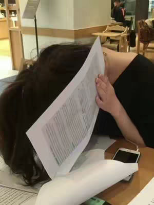The image shows a person, appearing to be a student, with their head resting on a desk covered in papers. The person's dark hair obscures their face as they lean against the pages. A smartphone placed on the desk next to them and in the background, a person is seated at a table.