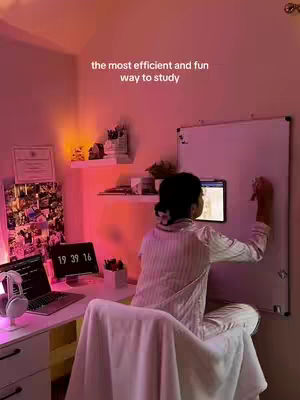 A person is facing away from the camera in a room with pink and orange lighting, leaning on a white desk. They're writing on a whiteboard. Behind them is a laptop, and a certificate is on the wall. The room shows a well-lit study setup.