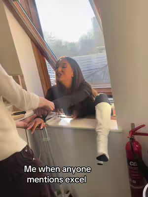A woman is seated in a window frame, with one leg extended out a window. She is leaning forward and gesturing with her hands, as if in conversation. Text at the bottom says, 'Me when anyone mentions excel'. There is a fire extinguisher in the background.