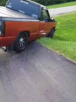 A side view of an orange and dark gray truck is pictured on a paved road, with one of the truck's tires partially off the road and onto a grassy area.