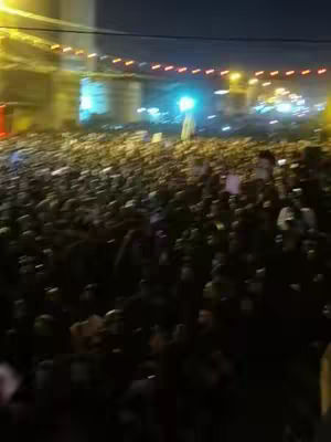 A low-angle, slightly blurred shot of a large crowd gathered at night, likely in an outdoor event. The crowd appears dense, filling the frame. There are lights visible in the background, possibly from buildings or a bridge, with various light sources illuminating the scene and the people within it.