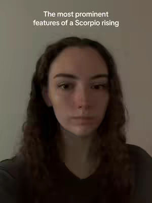 A medium shot of a young woman with long, wavy brown hair posing for a selfie. She is looking directly at the camera, her expression neutral. The background is a plain, light-colored wall. Text at the top of the image reads: 'The most prominent features of a Scorpio rising'.