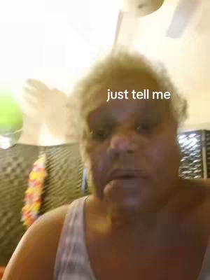 A selfie-style shot of a person with unkempt, light-colored hair. The person has a neutral expression and is centered in the frame. The background is out of focus and appears to be a room lit by a single light source.