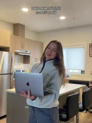 A woman stands in a kitchen holding a laptop. The kitchen has light-colored cabinets and a large island with bar stools. The woman is wearing a light-colored fleece jacket and gray pants. The overall composition is well-lit and inviting.