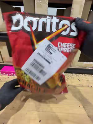 A person wearing black gloves is holding a bag of Doritos Cheese Pretzel crisps. The bag is predominantly red with white Doritos branding. A white address label is affixed to the bag. The background includes a wooden surface and cardboard boxes.