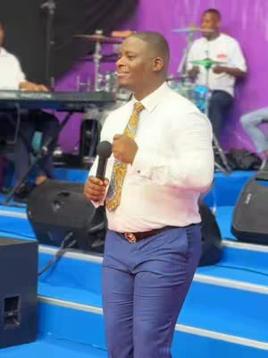 A man in a white shirt and blue pants is holding a microphone, smiling, and addressing a crowd. He is wearing a patterned tie. Behind him, other people are on the stage, with musical equipment.