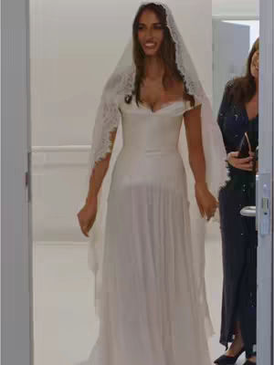 A woman in a white wedding dress and veil walks through a doorway, smiling. Another woman stands behind her. The scene is brightly lit, with a clean, modern aesthetic.