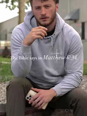 A man in a light gray hoodie sits outdoors, holding a smartphone. Text overlay reads, 'The Bible says in Matthew 6:34'. The background is blurred with greenery, creating a soft, natural ambiance. Overall, the image conveys a message of faith with the man posing in a relaxed manner.