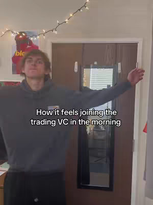 A young man is standing in a room with his arms outstretched. He is wearing a gray hoodie and dark pants. Text overlayed on the image reads, 'How it feels joining the trading VC in the morning.' The background includes a door with a mirror, a poster, fairy lights, and other interior elements.