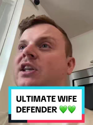 A man with short auburn hair and blue eyes is looking slightly up and left, mouth open as if speaking. He is in an indoor setting with a white wall behind him. The image includes a text box at the bottom with the words 'ULTIMATE WIFE DEFENDER' and two green heart emojis.