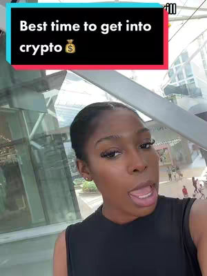A woman with dark hair is in a selfie video. She is wearing a black top and standing in front of a large, glass building. A text overlay above her head reads, "Best time to get into crypto" against a blacked-out rectangle, and there's a money bag icon next to the word crypto. The background has the blurry features, with a walkway and other people are visible in the background.