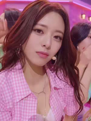 Close-up shot featuring a young woman with long, dark hair and pink checkered shirt. She has prominent brow and striking eyelashes, and is looking directly at the camera with a slight, subtle expression. The background is blurred, suggesting a stage or performance setting.