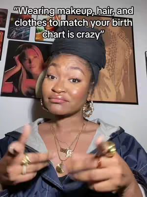 A woman with dark skin wearing a black headscarf, gold jewelry, and a navy blue jacket gestures with both hands in a selfie-style shot, against a backdrop of framed artwork. The text overlay reads "Wearing makeup, hair, and clothes to match your birth chart is crazy". The overall composition suggests self-expression and commentary on astrology.