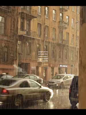 An image of a city street in the rain. Cars are driving on the wet street, and rain streaks down the image. There are buildings in the background.