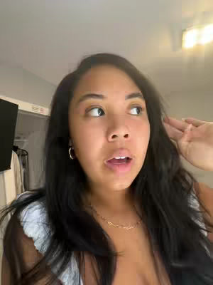 A woman with dark hair and fair skin looks off to the side with a surprised expression. She is wearing gold earrings and a gold necklace. The background is a basic room with lights and a closet.