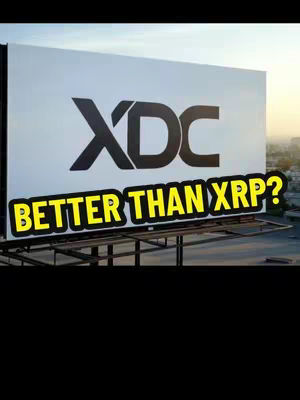 A daytime shot of a billboard with the XDC logo in large, bold black text over a white background. Below, in a yellow font with a black outline, the text 'BETTER THAN XRP?' is prominently displayed. The billboard sits outside with a city in the background.