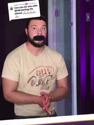 A man with short brown hair and a beard stands in front of a purple and white background. He has a black bar over his mouth. Above his head is a white speech bubble with text in it.
