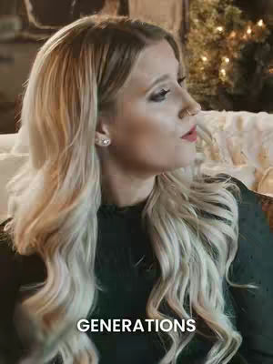 A woman with long blonde hair, wearing a black top, is seen in a medium shot. She has makeup on, with a slight smile and is looking slightly off to the side. The background behind her is softly lit with what appears to be a Christmas tree and cozy decor. The text "GENERATIONS" is overlaid in white at the bottom.