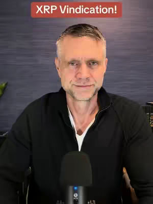 A man with short gray hair and stubble is centered in the frame, facing the camera with a slight smile. He wears a black zip-up jacket over a white collar and is positioned in front of a dark blue background. Below him, a microphone is visible. A red banner with white text is at the top.