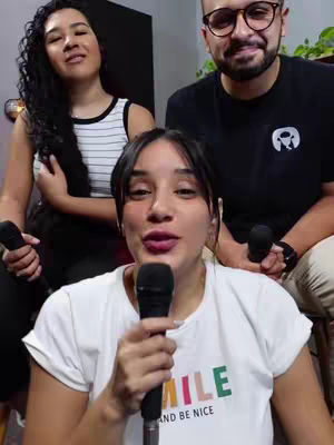 A cover image of three people in a room setting seemingly recording a video. The main subject in the foreground is a woman holding a microphone wearing a white shirt with a colorful logo. The other two people are in the background and holding a microphone, one is a woman and the other is a man.