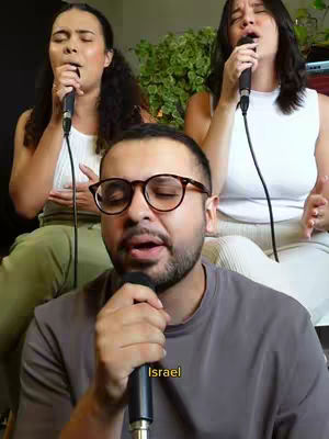 The cover image features three people: a man in the foreground wearing glasses, singing into a microphone, with two women in the background, also singing into microphones. The composition is a medium shot, focusing on the singers, with a neutral background. The color palette is natural, focusing on skin tones and the lighting is well distributed.