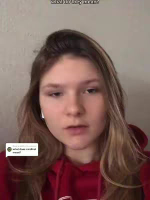 A young woman with fair skin and long blonde hair is centered in the frame. She wears a reddish-burgundy hoodie and appears to be speaking. A small, white speech bubble with text sits in the lower left corner. The background is blurred, but appears to be a light-colored wall.
