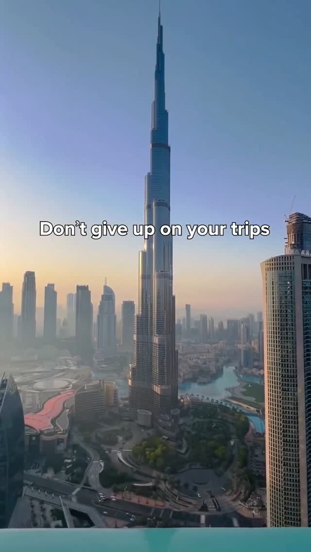 An aerial view of a cityscape with a focus on the Burj Khalifa, a tall, slender skyscraper that dominates the center of the frame. Surrounding it are other tall buildings, a river or body of water, and overall, a clear sky. The text overlay, in a white, sans-serif font, states, "Don't give up on your trips."