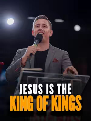 A man in a gray blazer and black shirt speaking into a microphone in front of a clear lectern. Text overlayed on the image reads: JESUS IS THE KING OF KINGS.