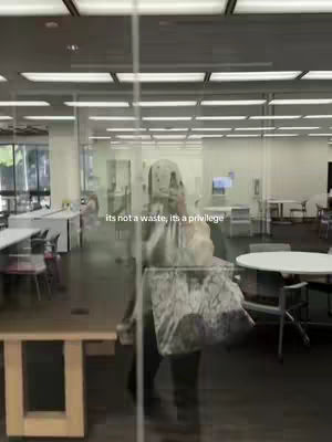 A person is reflected in a glass pane, appearing to be in a professional or educational setting like a library or office. They are wearing a patterned bag, which is partially visible.