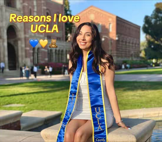 A woman wearing a gown with UCLA written on is sitting on a stone bench with grass and buildings in the background. The woman is smiling towards the camera. The words "Reasons I love UCLA" are written in a yellow font at the top, along with a blue heart, yellow heart and bear emojis arranged to the left of the woman.
