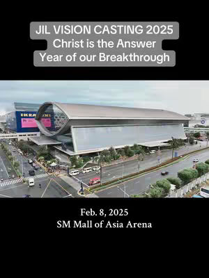 An aerial view of a modern, multi-story building with a rounded glass window at the front. The image is dominated by the building, with a black bar at the bottom containing descriptive text. Above the building is a gray banner containing text. The setting appears to be an urban environment with roads and other buildings visible.