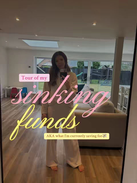 A woman stands in a modern room, appearing to take a selfie in a large mirror. She is the main subject, dressed in a neutral-colored outfit. Text overlays with playful fonts highlight the content's themes: "Tour of my" in a pink rectangle bubble, and "sinking funds" in large, layered yellow script. A smaller text bubble states "AKA what I'm currently saving for". The house interior presents a muted palette, creating a clear focal point on the individual.
