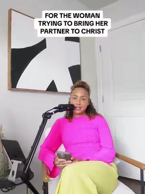 A woman in a bright pink top and yellow pants sits in a white chair, holding a phone and looking at the camera. A microphone stand is on her left and a piece of abstract art is framed behind her head.