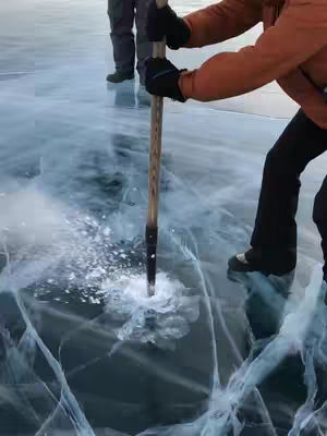 A person is seen using a tool to break through thick ice. The lighting is natural, and the scene is set outdoors. The composition is straightforward, with the person focused on their task with someone else in the background.