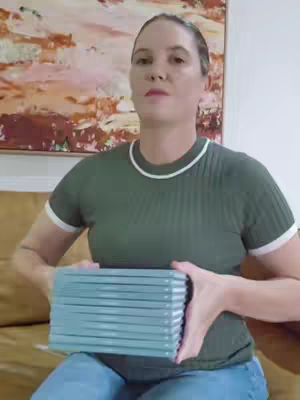 A woman in a green shirt and blue jeans sits holding a stack of books. She is indoors, with an abstract painting on the wall behind her.