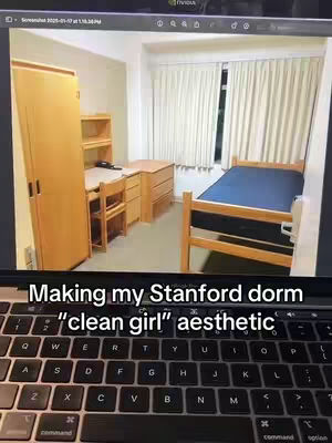 A dorm room is shown in a rectangular frame, with a laptop keyboard showing to the bottom of the frame. The dorm has a bed with blue sheets, a wooden desk, a dresser, and a wardrobe. The lighting is natural from a window with closed curtains.