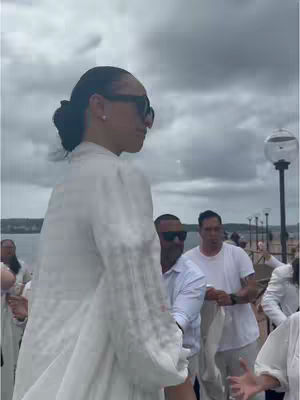 A woman wearing sunglasses and a white dress stands facing left, with a group of people in white clothing behind her. The background suggests an outdoor setting with a body of water and a cloudy sky.