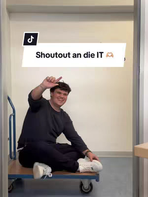 A young man sits cross-legged on a small wheeled cart, raising his hand in a greeting or salute. He is smiling and appears to be inside a building, likely a hallway or storage area. The background is a plain, light-colored wall.