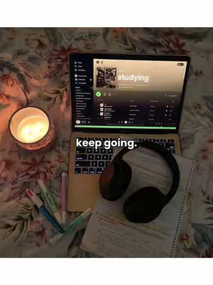 The image shows a laptop open on a bed covered with a floral blanket. A candle is lit to the left of the laptop, and there are highlighters and pens scattered around the base. The laptop screen displays a music app with the word "studying" at the top and the words "keep going."