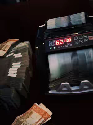 The image shows a money-counting machine with a digital display showing '8862 100.' There are stacks of cash on either side, with some currency visible. The scene is dark, and the money is the central focus.