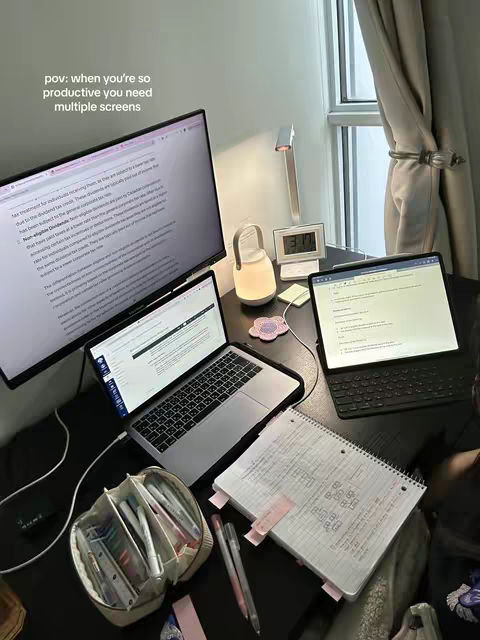 A well-lit desk with multiple screens and study materials, including a laptop, notebook, pens, and a pencil case. The composition is tidy and suggests a productive study environment. Soft light comes from a lamp and a digital clock is shown.