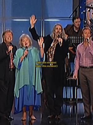 A group of five people on a stage, mid-performance. The lighting is dim, with a blue backdrop. Several individuals are holding microphones singing, while one person enthusiastically raises their arm, holding a microphone. A text overlay includes the phrase take this moment.